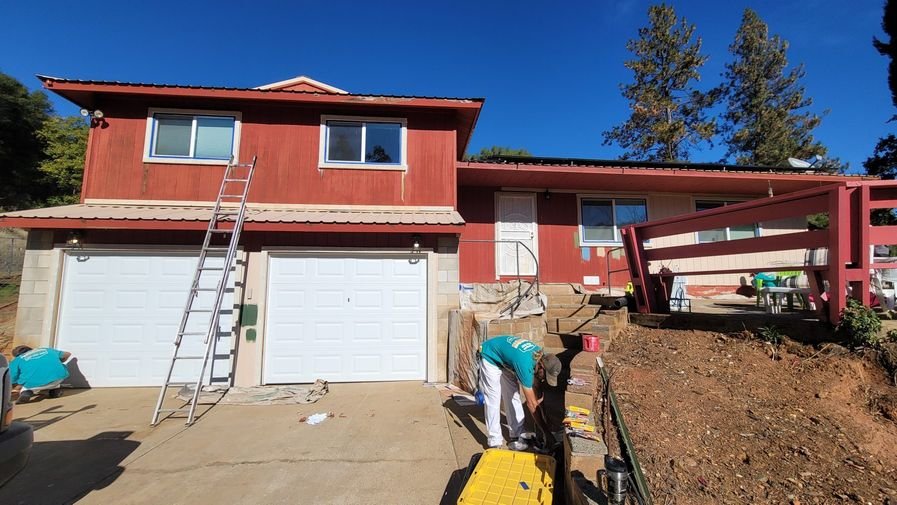 A weather-worn red house mid-prep, siding faded, a ladder up and a crew member scraping before painting