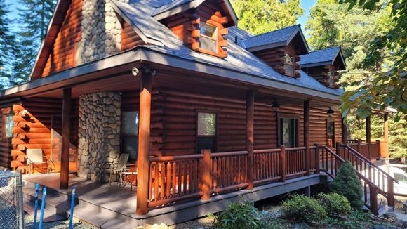 A log home with a wraparound porch, freshly stained a rich amber, framed by pines