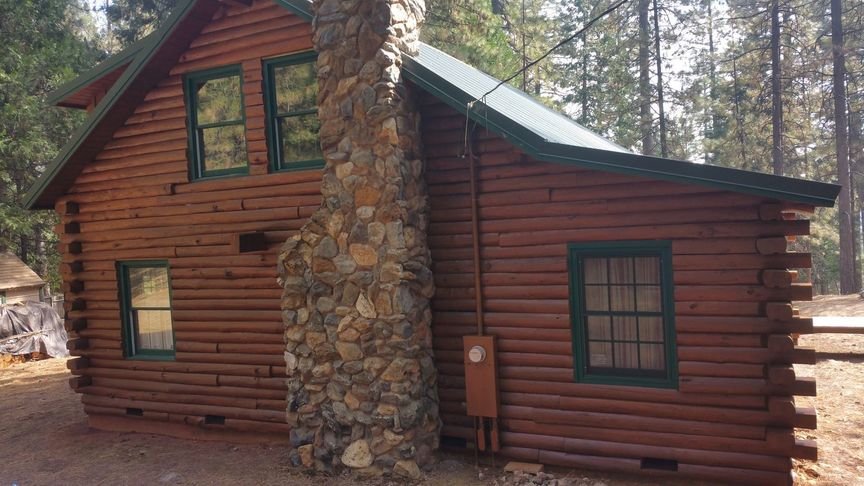 A tall amber-stained log home with a full stone chimney and green window trim