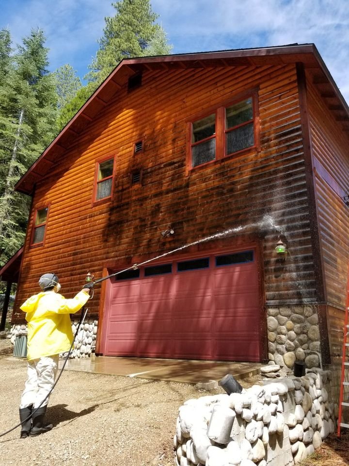 A Michael Hines Painting crew member in protective gear washing a log wall with cleaner to strip mildew before staining
